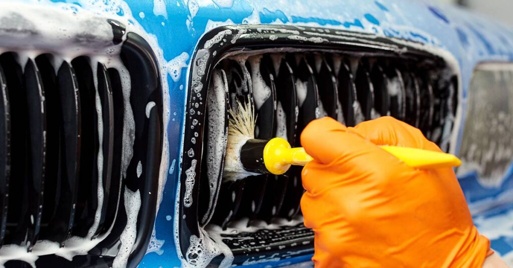 A hand in an orange glove cleans the front of a blue car with a yellow cleaning brush. The soap covers the car's front end.