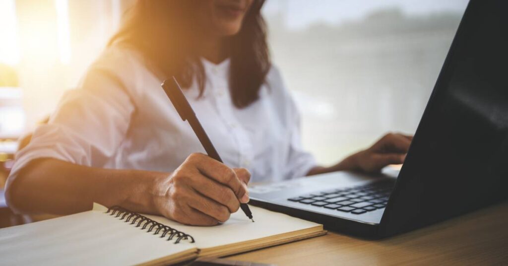 While researching online, a young woman with dark hair writes in a notepad. She's wearing a white button-down shirt.