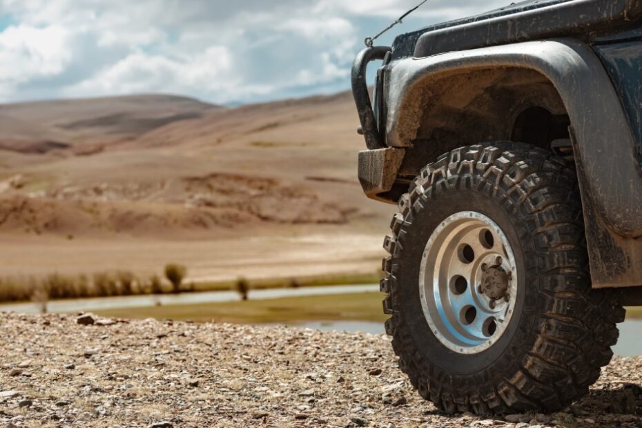 A black off-road vehicle drives down a gravel road bordering a lake. You can see a mountainous terrain in the distance.